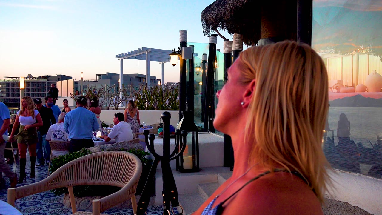 People move through the crowded terrace while the blond woman stays in the right foreground, with seated guests, glass wind screens, lamps, and sunset color visible in Cabo San Lucas, Mexico.