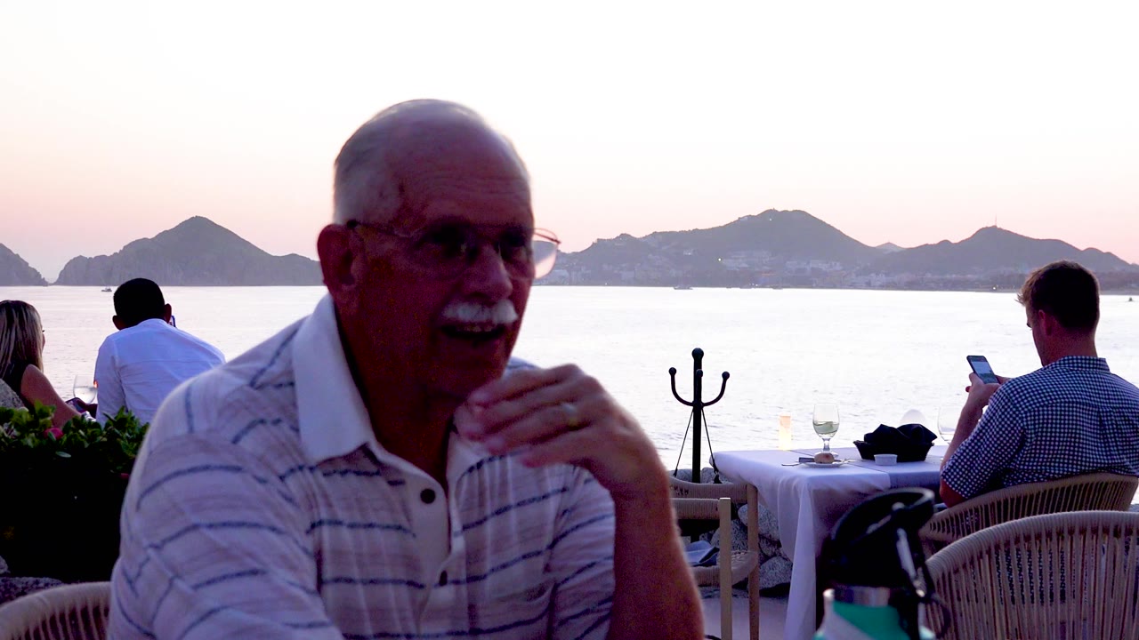 Older man at outdoor restaurant table with waterfront diners behind him.