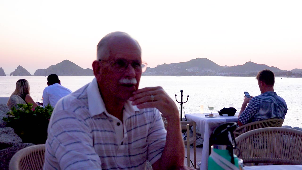 An older man in glasses rests his hand near his face at a Cabo San Lucas seaside restaurant, with a teal bottle, tableware, wicker chairs, diners, water, and low hills visible.