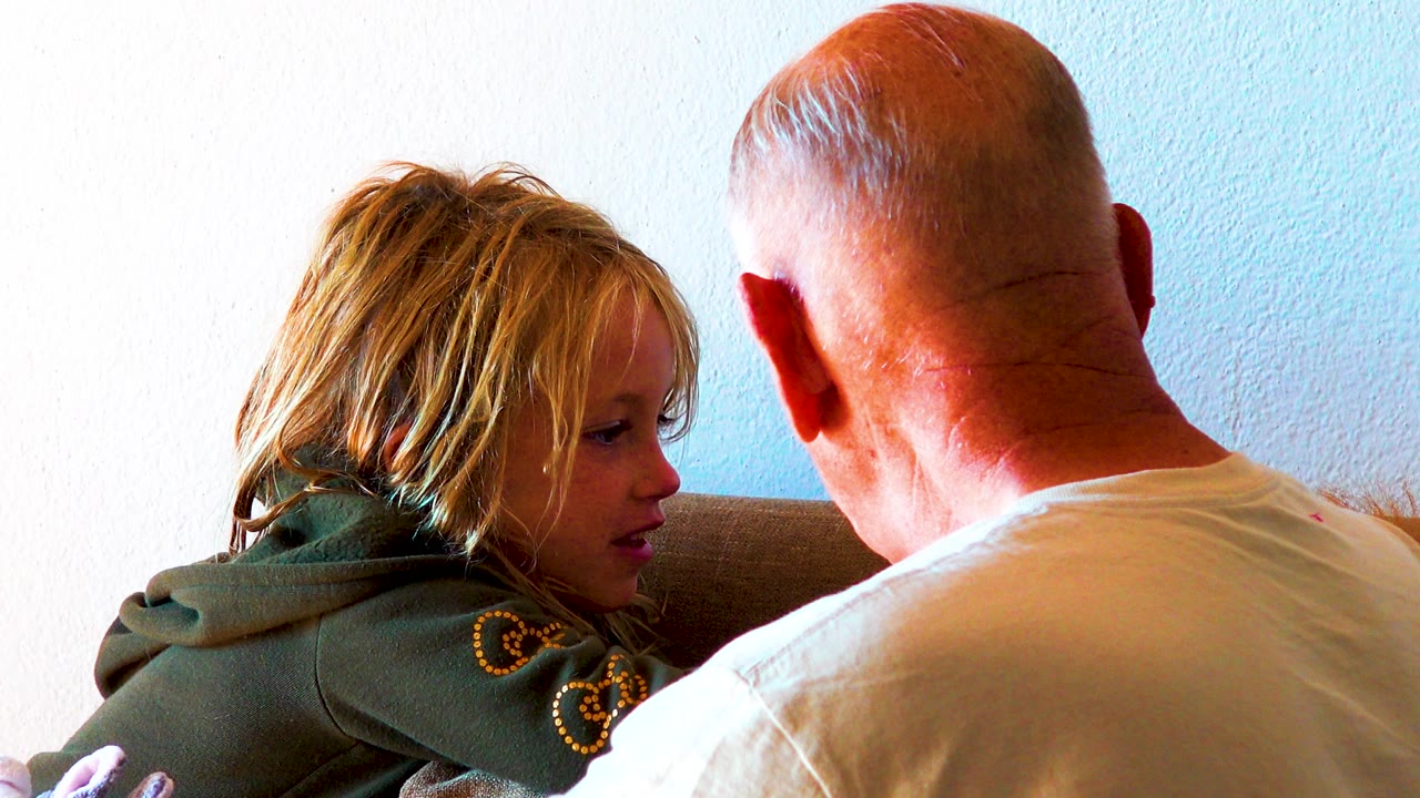 At the start of the Los Cabos clip, a blonde child in a green hoodie sits on a couch facing a bald adult man whose back fills the right side of the frame.
