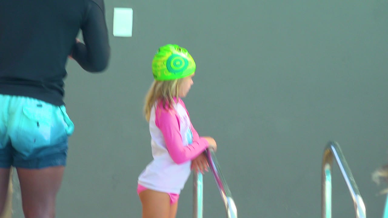 At 0:00, a child in a bright green swim cap stands beside a metal pool ladder in Cabo San Lucas, with a muted green wall behind and an adult partly visible at left.