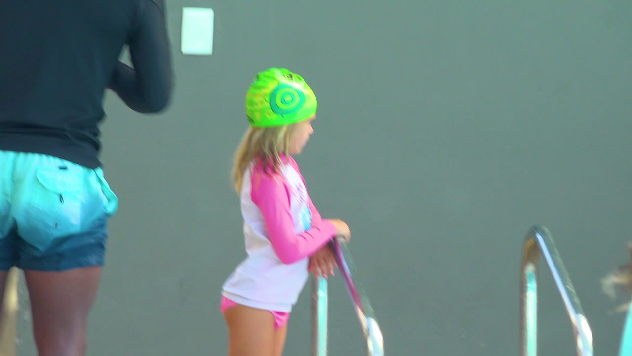 At 0:01, the child remains near the pool ladder in Cabo San Lucas while a cropped adult in blue shorts occupies the left foreground against the plain wall.