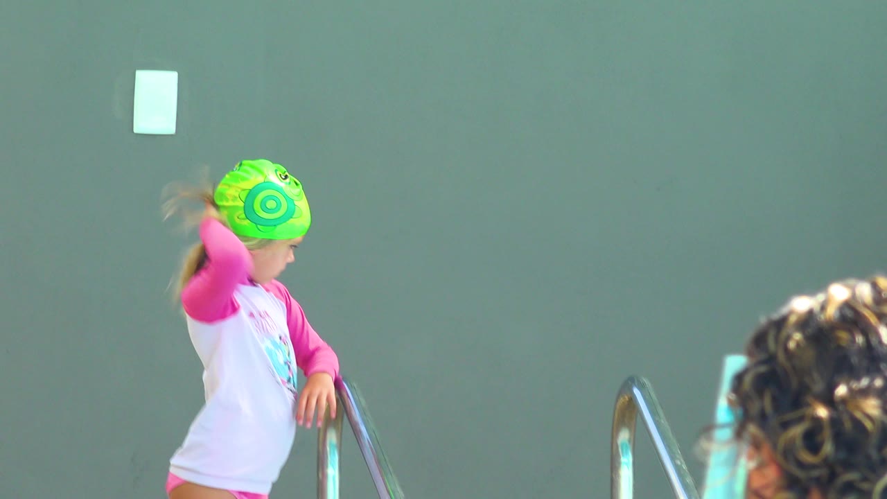 At 0:02, the child reaches toward the green swim cap beside the pool ladder, with the wall and a blurred adult head at the lower right of the Cabo San Lucas pool area.