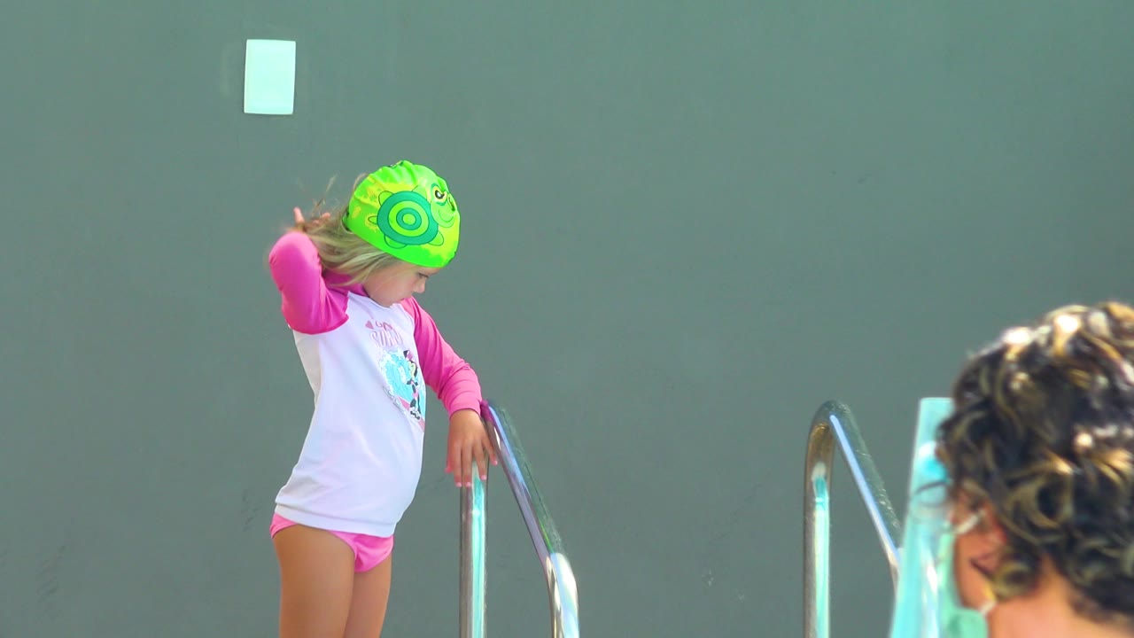 Child at pool ladder with towels and masked adult partly visible.