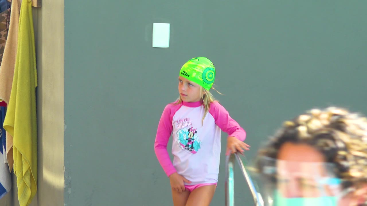 In Los Cabos, Mexico, the girl stands slightly farther right with one hand near the pool rail, while a cropped adult face and shoulder blur through the lower foreground.