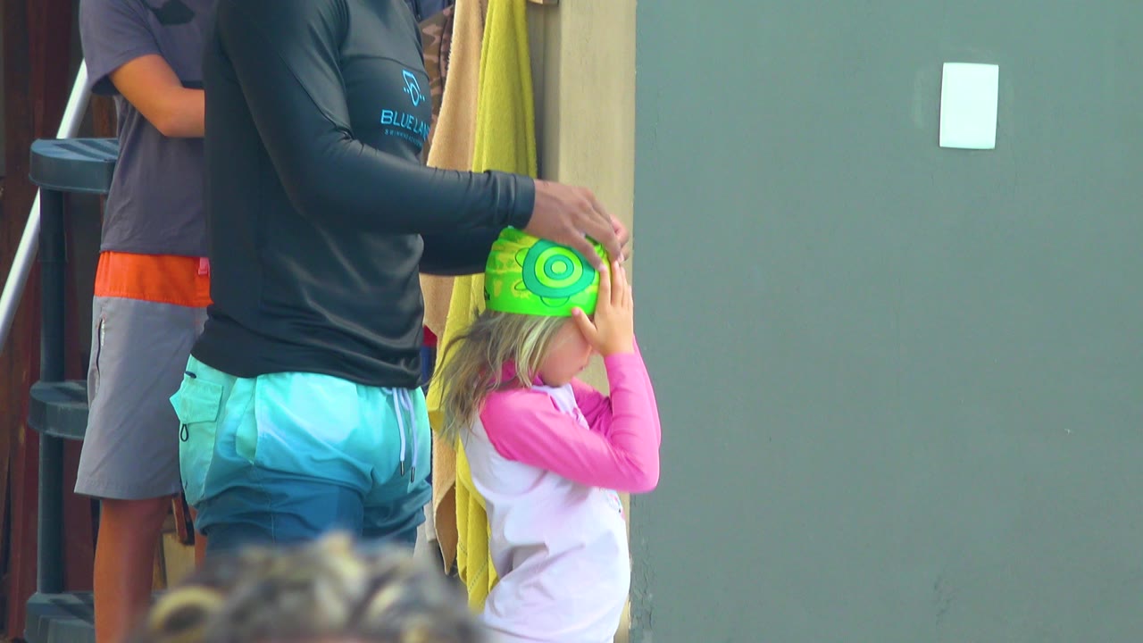 In Cabo San Lucas, Mexico, the child keeps both hands near the bright green swim cap while the adult steadies it next to hanging towels and a plain green wall.