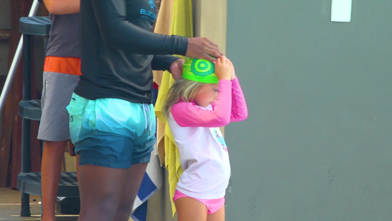 In Cabo San Lucas, Mexico, the child is seen in profile with hands on the bright green swim cap as the adult's hand begins lifting it away from the head.
