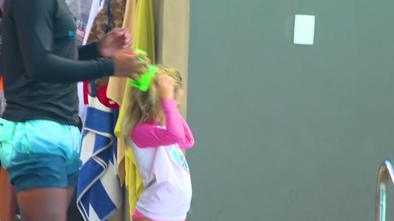 In Cabo San Lucas, an adult in turquoise swim shorts adjusts a blonde child's hair beside hanging towels and a green wall.