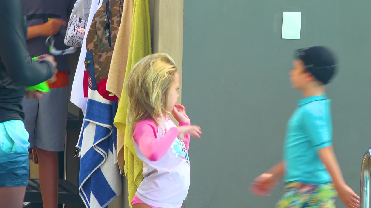 In Cabo San Lucas, the blonde child stands near the towels while the adult is partly out of frame and background figures appear near the wall.