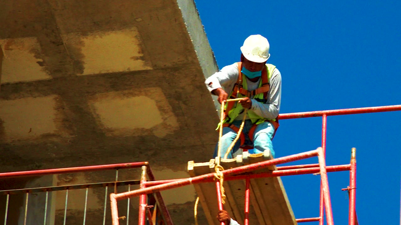 In Cabo San Lucas, Mexico, a construction worker in a white hard hat and safety vest leans over red scaffolding beneath a concrete overhang at the start of the clip.