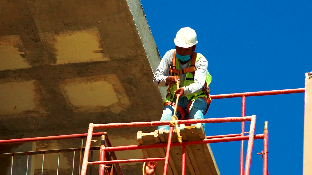 In Cabo San Lucas, Mexico, the worker looks down while pulling or positioning the orange pipe beside the red scaffold rail, with concrete and blue sky behind him.