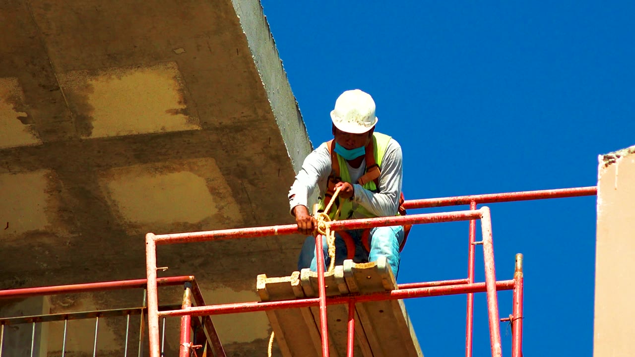 Worker leaning forward on scaffold with orange pipe in hand.