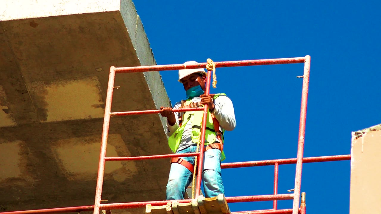 A low-angle view in Cabo San Lucas, Mexico shows the worker holding the red railing upright, with the safety harness visible over light clothing and clear blue sky behind.