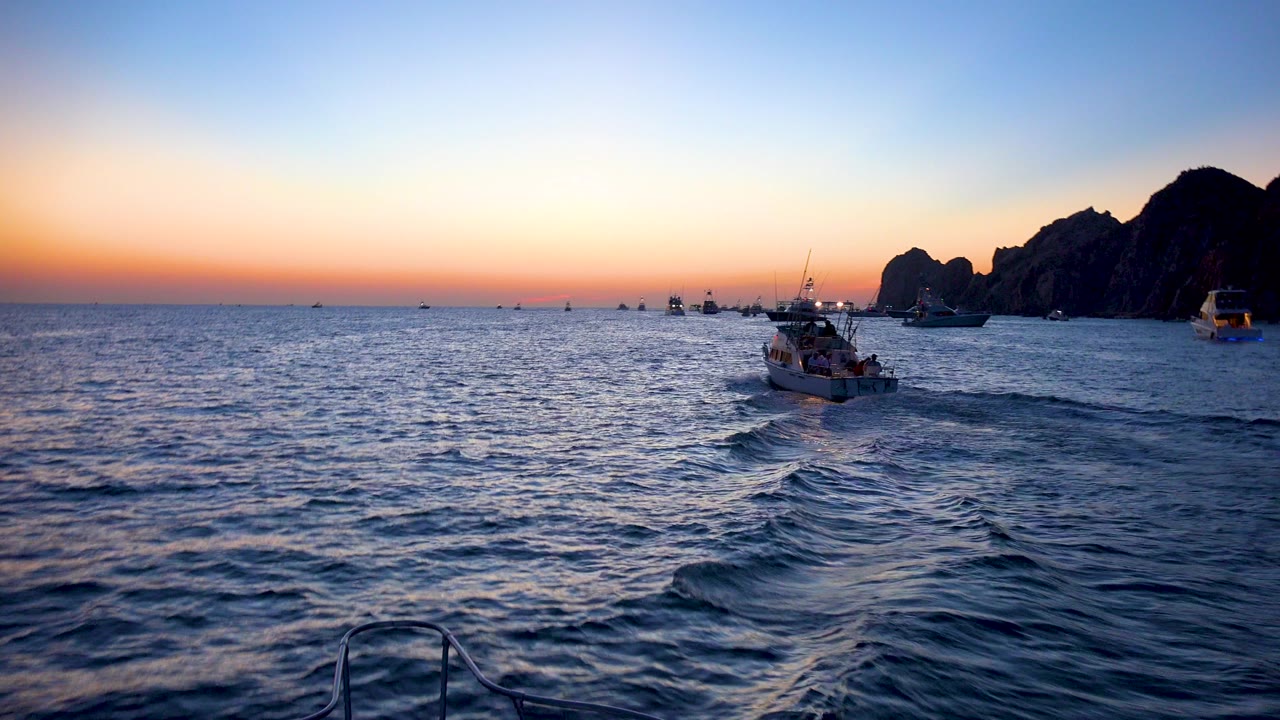 Cabo San Lucas ocean at sunset with a central small boat ahead, scattered distant boats, dark cliffs on the right, a low railing at the bottom edge, and no visible people.