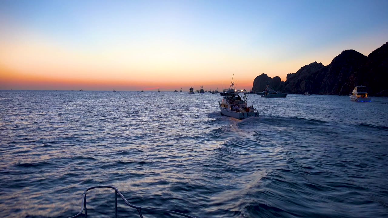 Cabo San Lucas coastline at dusk with the central boat slightly farther right, rippled blue water in the foreground, cliff silhouettes on the right, and no visible people.