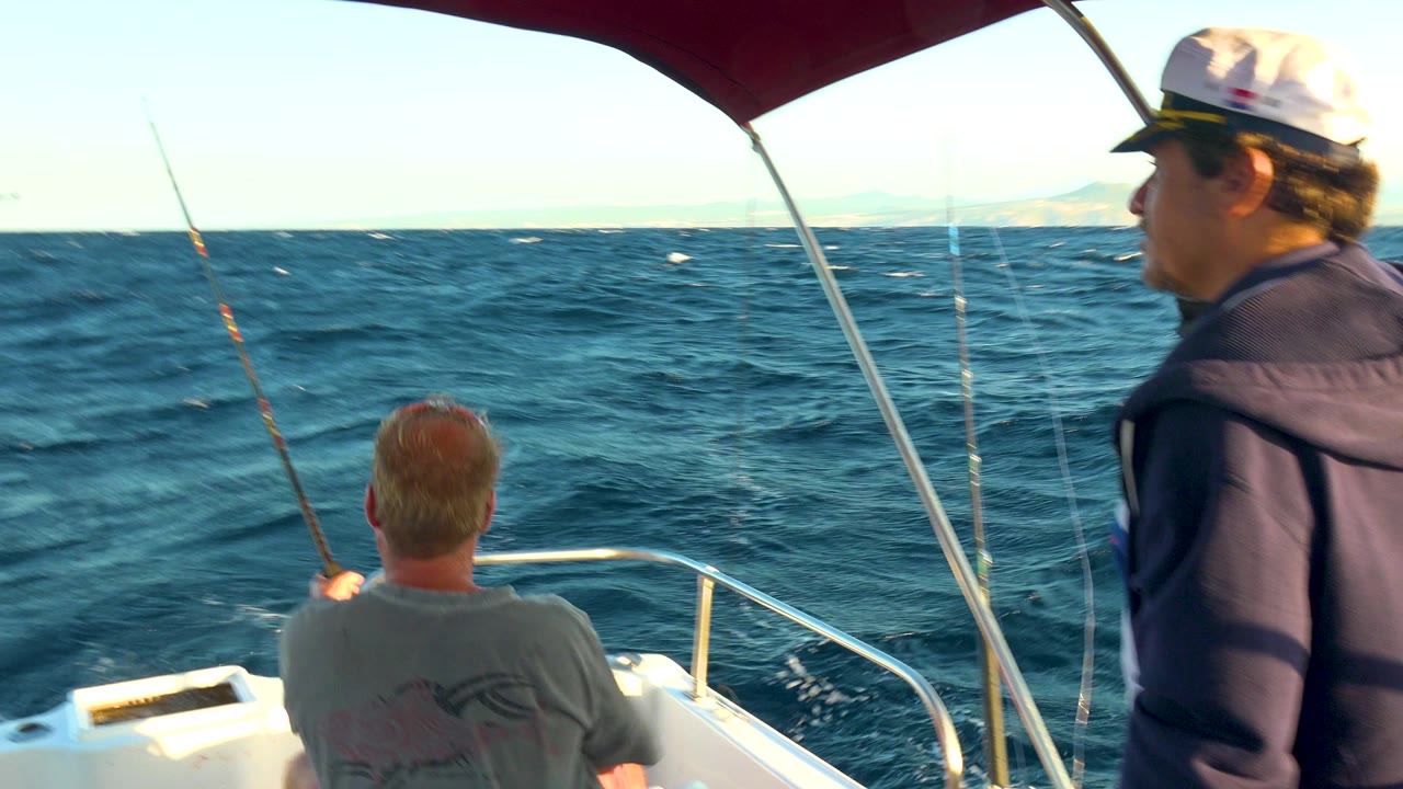 Standing man in cap and seated man look out from a boat toward open water.