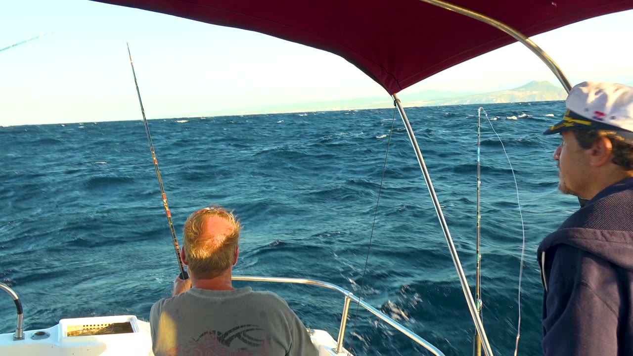 The seated passenger faces the choppy water in Cabo San Lucas, Mexico, while another man stands partly at the right edge beside upright fishing rods and boat rails.