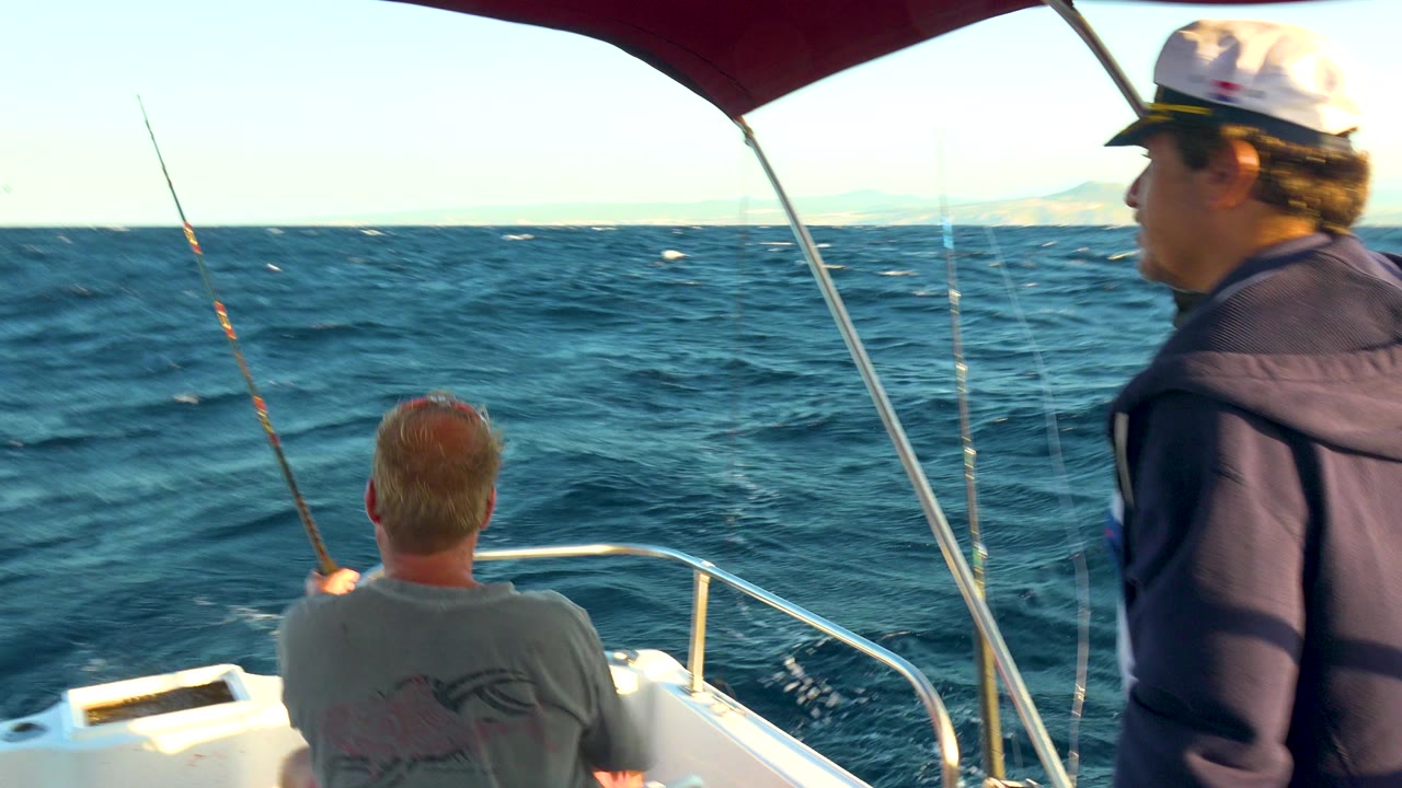 At 0:00, two adult men are aboard a small boat in Cabo San Lucas, Mexico, with one seated near the bow, one standing at the right edge, fishing rods upright, and choppy blue water ahead.