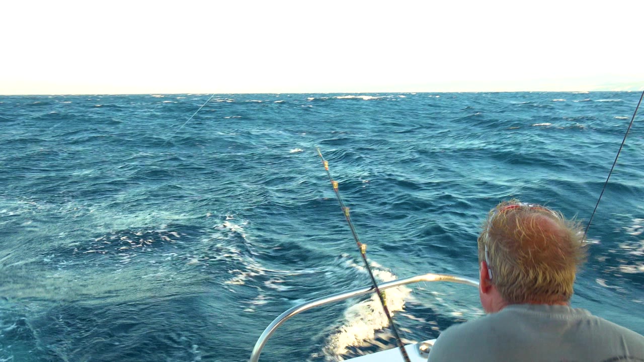 A man sits at the front edge of a boat near Cabo San Lucas, Mexico, facing choppy blue open water with a fishing rod rising through the foreground.