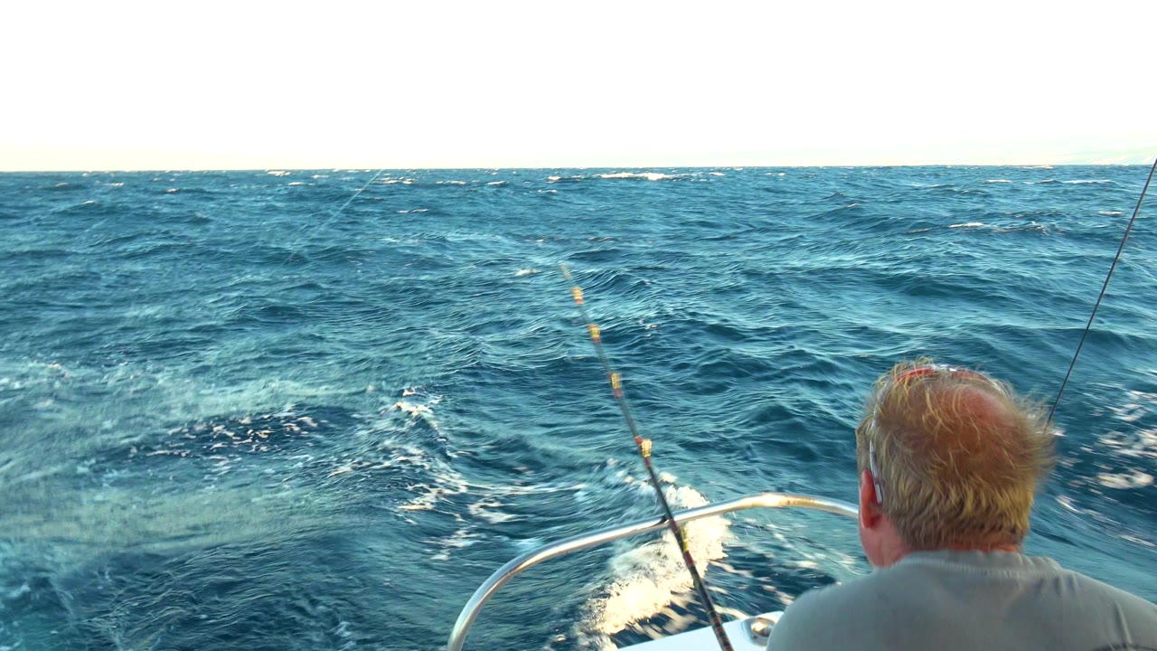Fishing rod and boat bow frame choppy blue water with a man seen from behind.