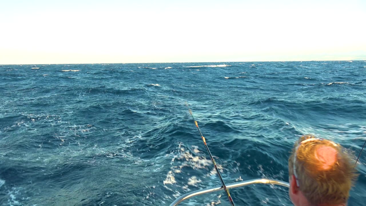A partial view of the man appears at the right edge while the boat rail and fishing rod cut across choppy Cabo San Lucas open water under bright light.