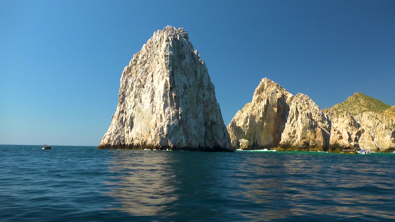 Opening frame of Cabo San Lucas rock formations above rippled blue ocean water, with bright sun on pale stone and no visible people.