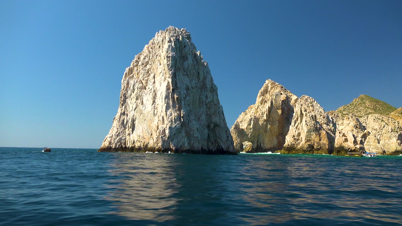 Tall sea rock with smaller rocky shoreline behind blue water.