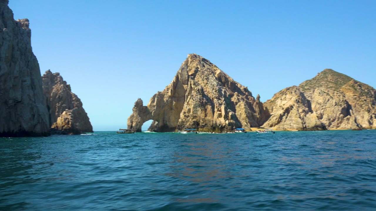 A tall shaded cliff edge enters at left while sunlit Cabo San Lucas rock formations, a sea arch, small boats, and blue water remain visible, with no people seen.