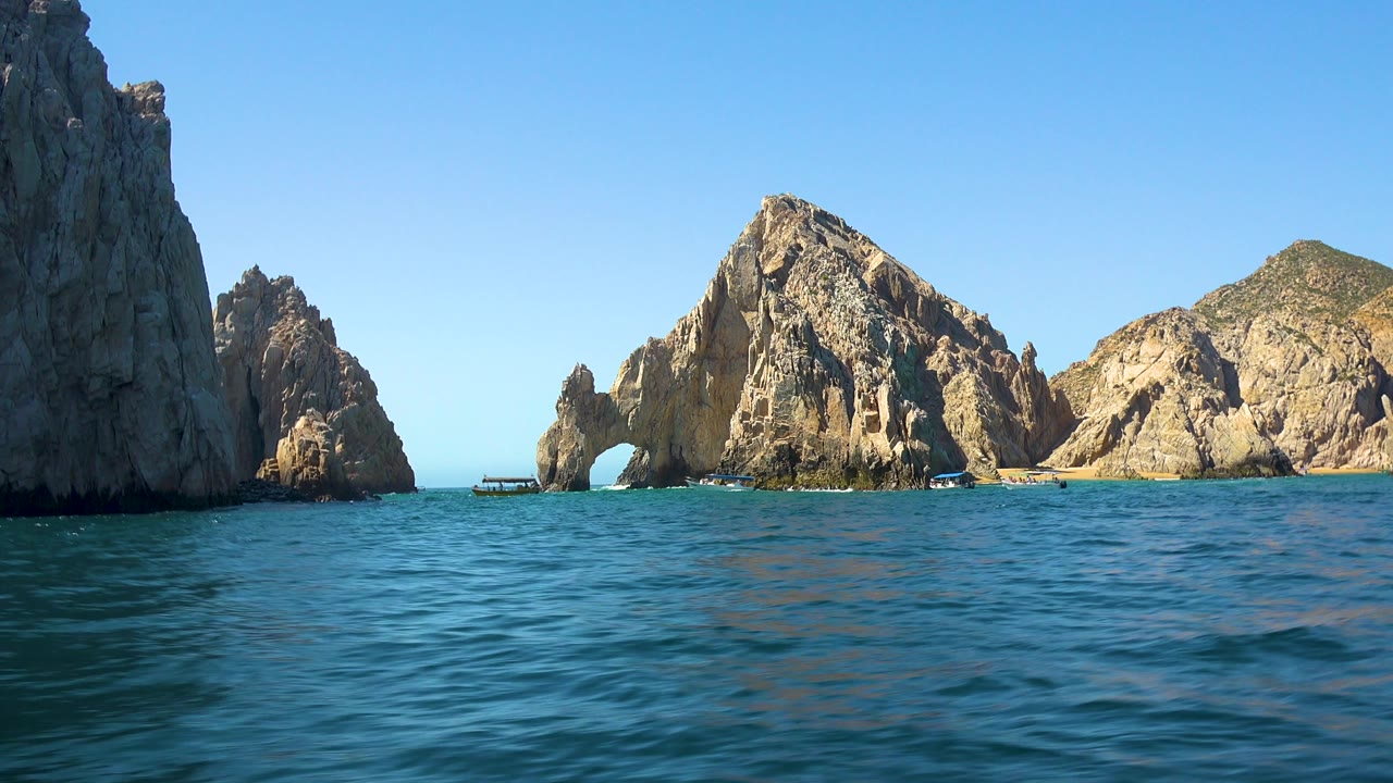 The frame shows a shaded rock wall on the left, the sea arch and larger tan cliffs across the water, small boats near the shoreline, and no visible people in Cabo San Lucas.