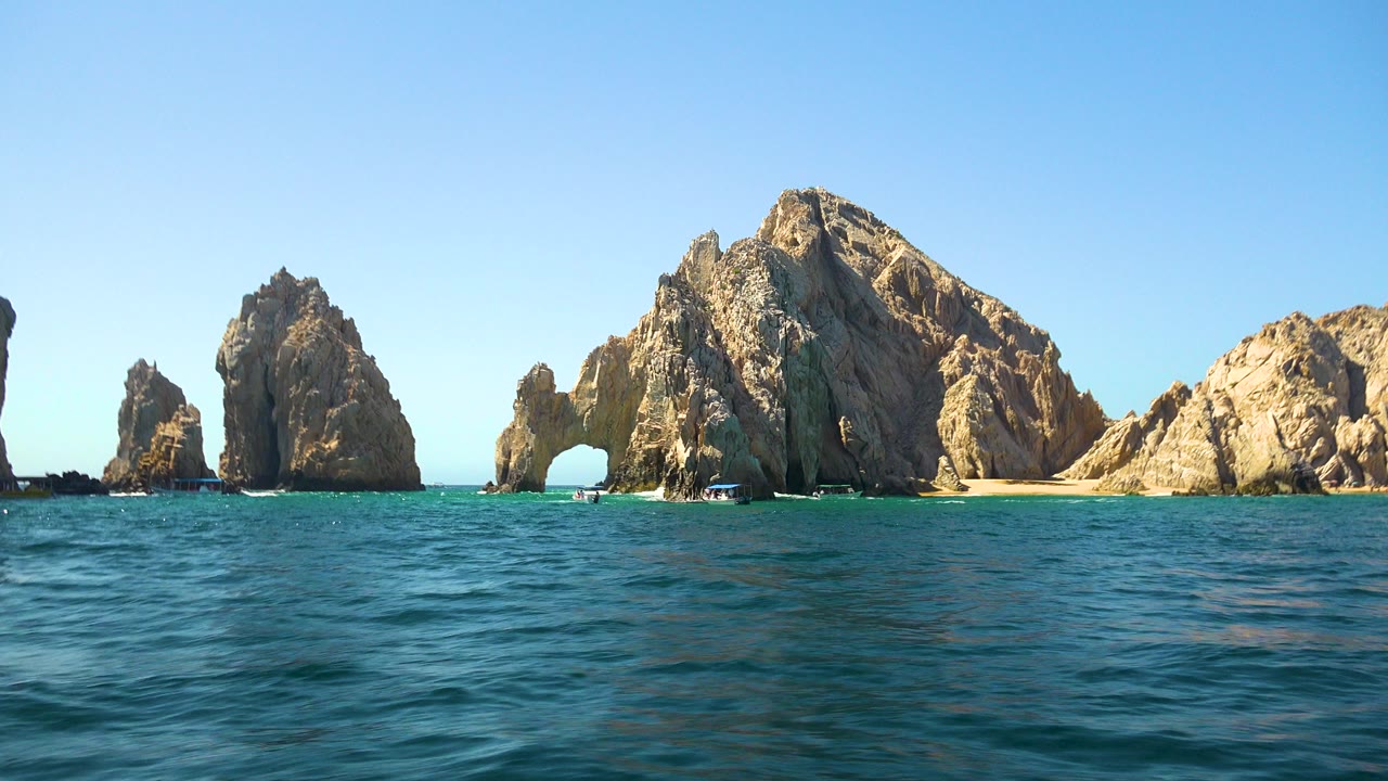 Wide Cabo San Lucas frame with rippled blue-green water in the foreground, tan rock formations and a natural arch across the middle distance, small boats near the rocks, and no visible people.
