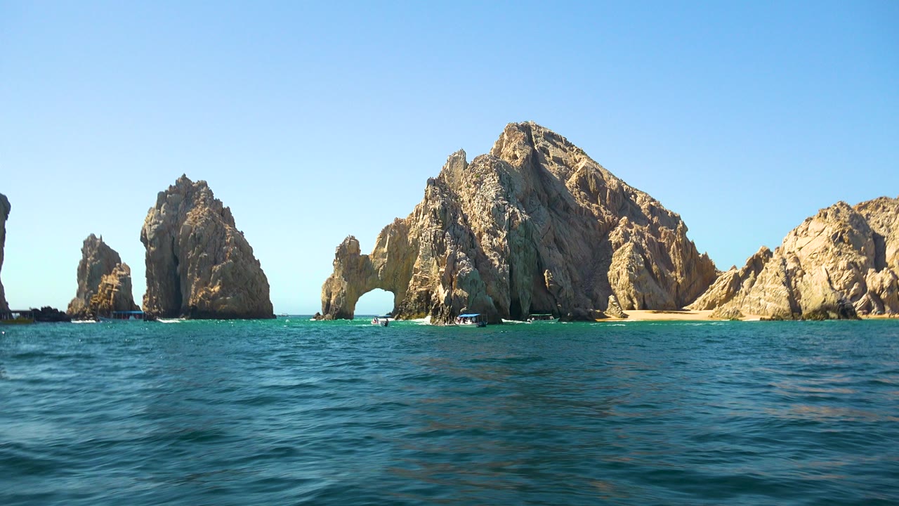 Cabo San Lucas view with the waterline low in frame, the natural arch left of the largest rock face, small boats near the base, and no visible people.