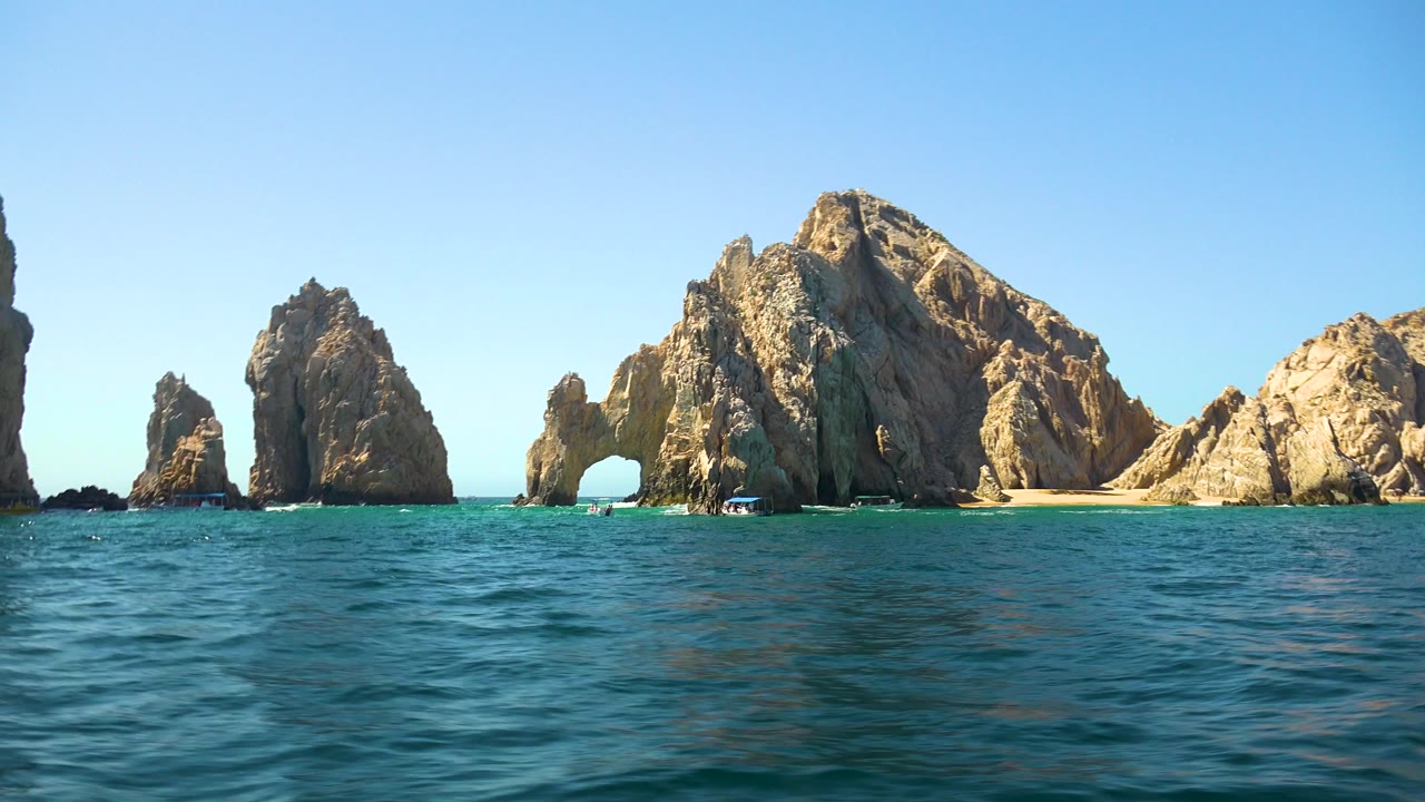 Cabo San Lucas frame with choppy foreground water, several separated rock stacks, the arch opening near center-left, a large sunlit rock mass to the right, and no visible people.