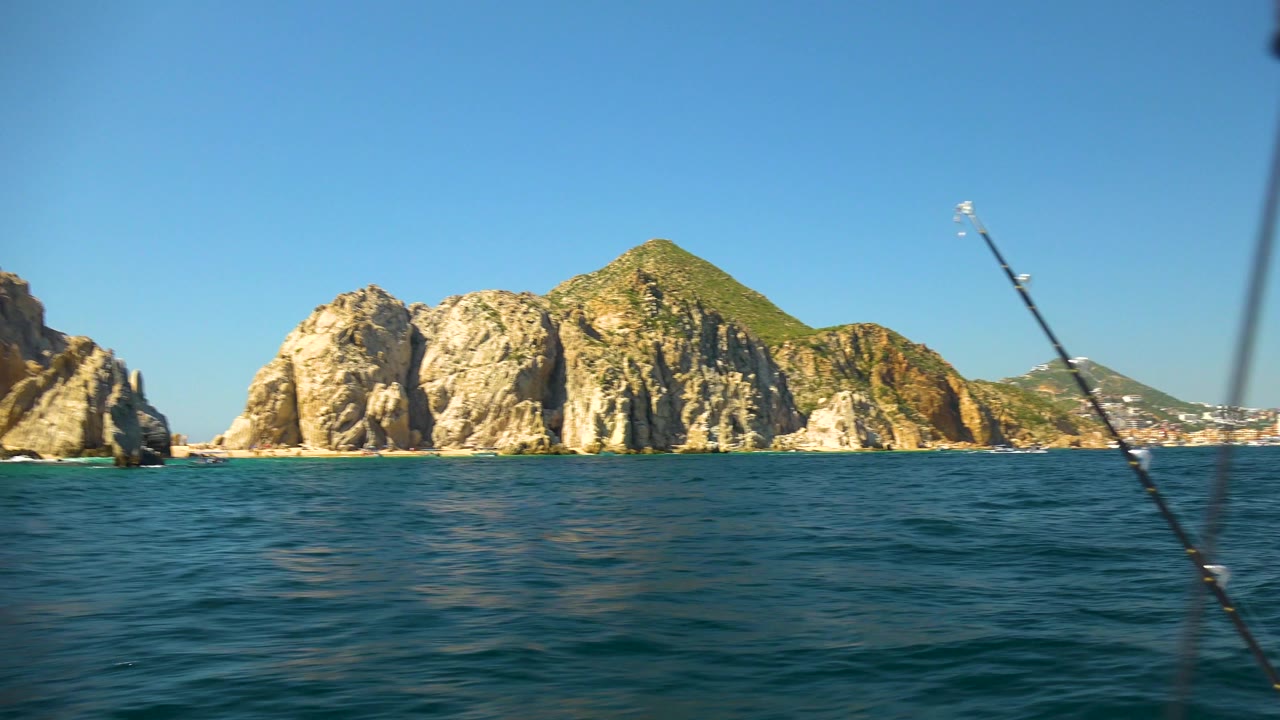 Cabo San Lucas rocks and hillside appear beyond the water while dark fishing rods cross the foreground, with no visible people.
