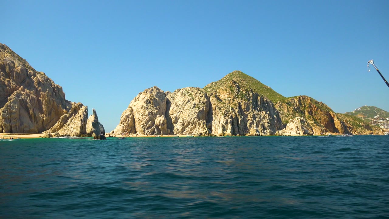 Wide boat-level view of blue water facing rocky cliffs and green hills in Cabo San Lucas, Mexico, with no visible people.