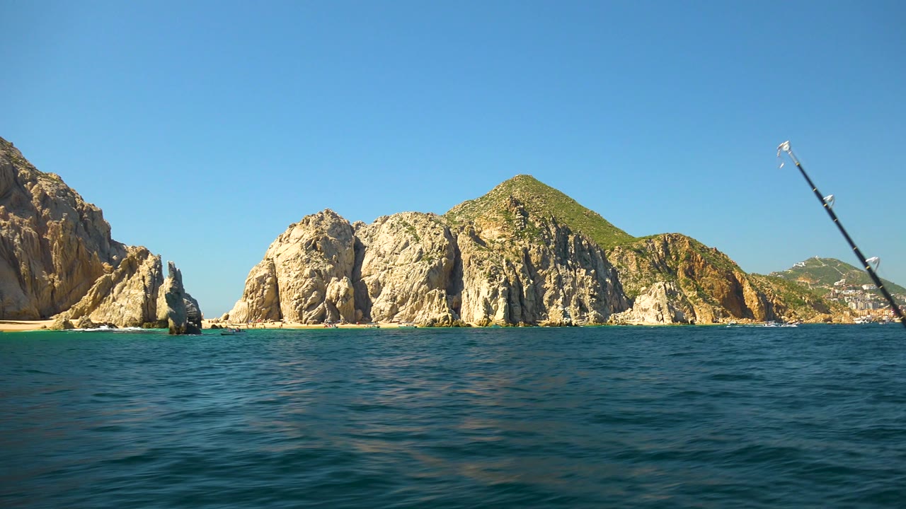 A fishing rod leans across the right side of a boat-level view toward rocky cliffs and green hills in Cabo San Lucas, Mexico, with no visible people.