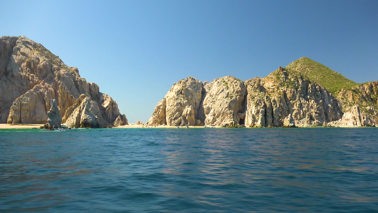 Cabo San Lucas rocky coastline seen across rippled blue ocean under clear daylight, with no visible people in the opening frame.