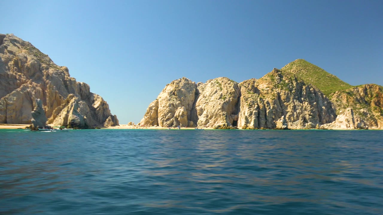 A wide view of Cabo San Lucas rock formations spans the frame across blue ocean, with bright sky overhead and no visible people.