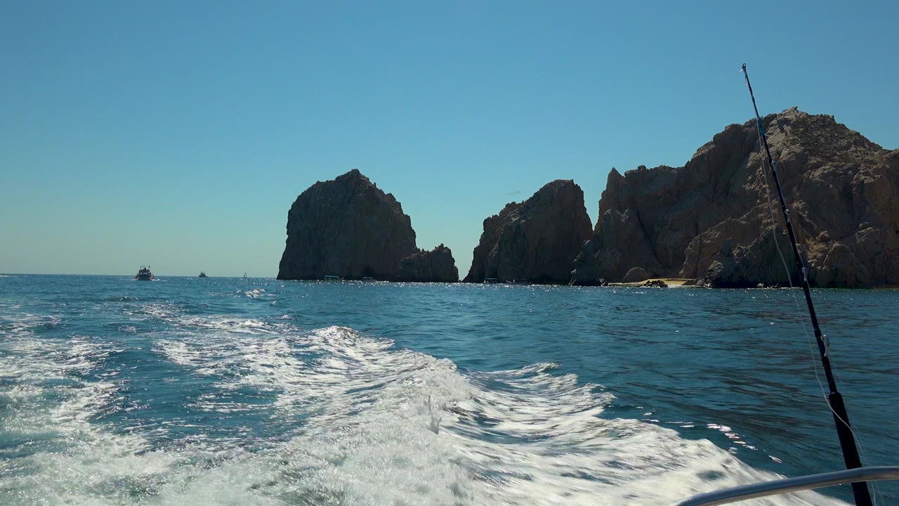 Cabo San Lucas ocean view from a boat, with white wake in the foreground, large rock formations across the horizon, a fishing rod at the right edge, and no visible people.