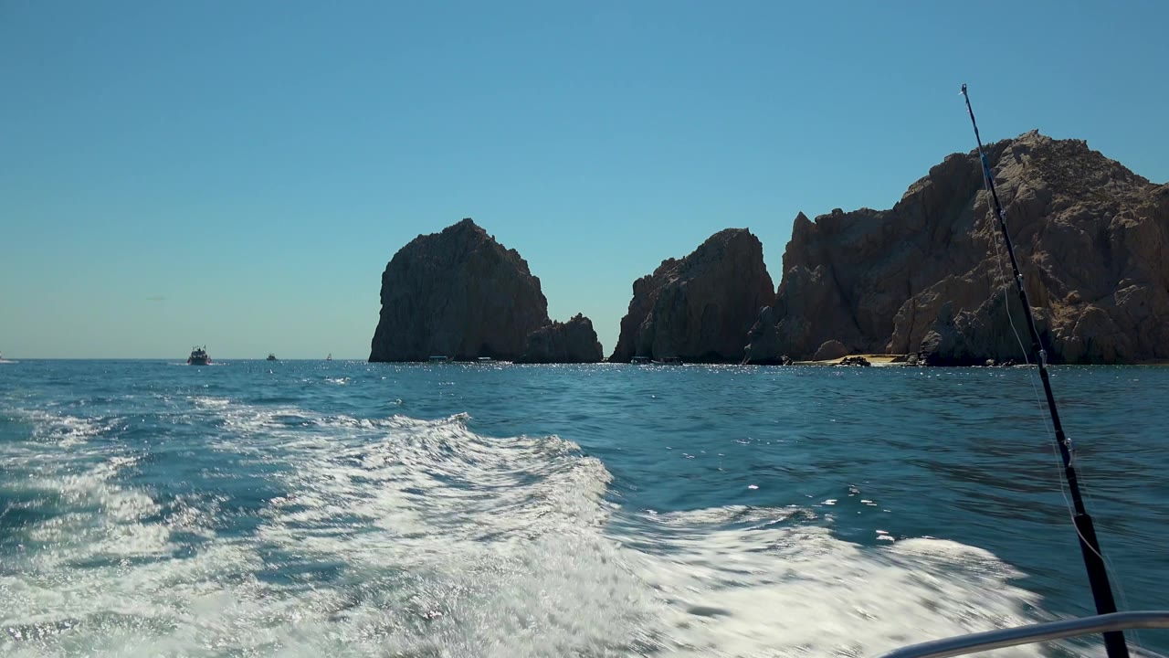 Cabo San Lucas frame with churning white wake across the lower half, rocky formations spanning the horizon, a fishing rod on the right side, distant boats, and no recognizable people visible.