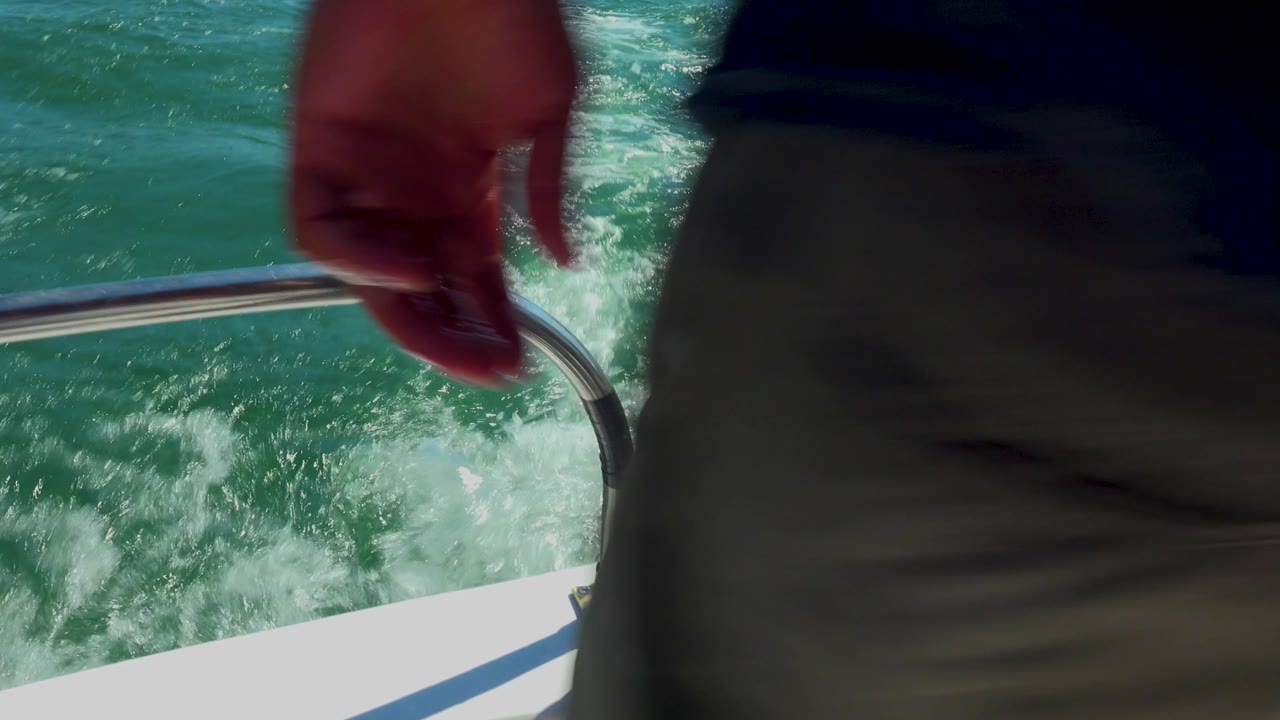 Partial human hand and torso fill the foreground beside a white boat rail over churning turquoise water in Cabo San Lucas, Mexico.