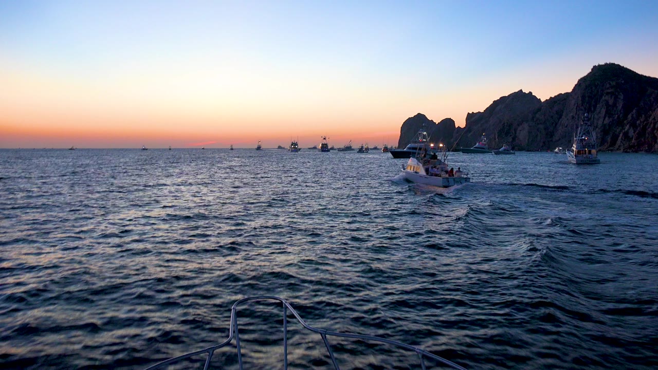 Cabo San Lucas, Mexico ocean view at dusk with small boats spread across the water, dark cliffs on the right, foreground boat railing, and no visible people.