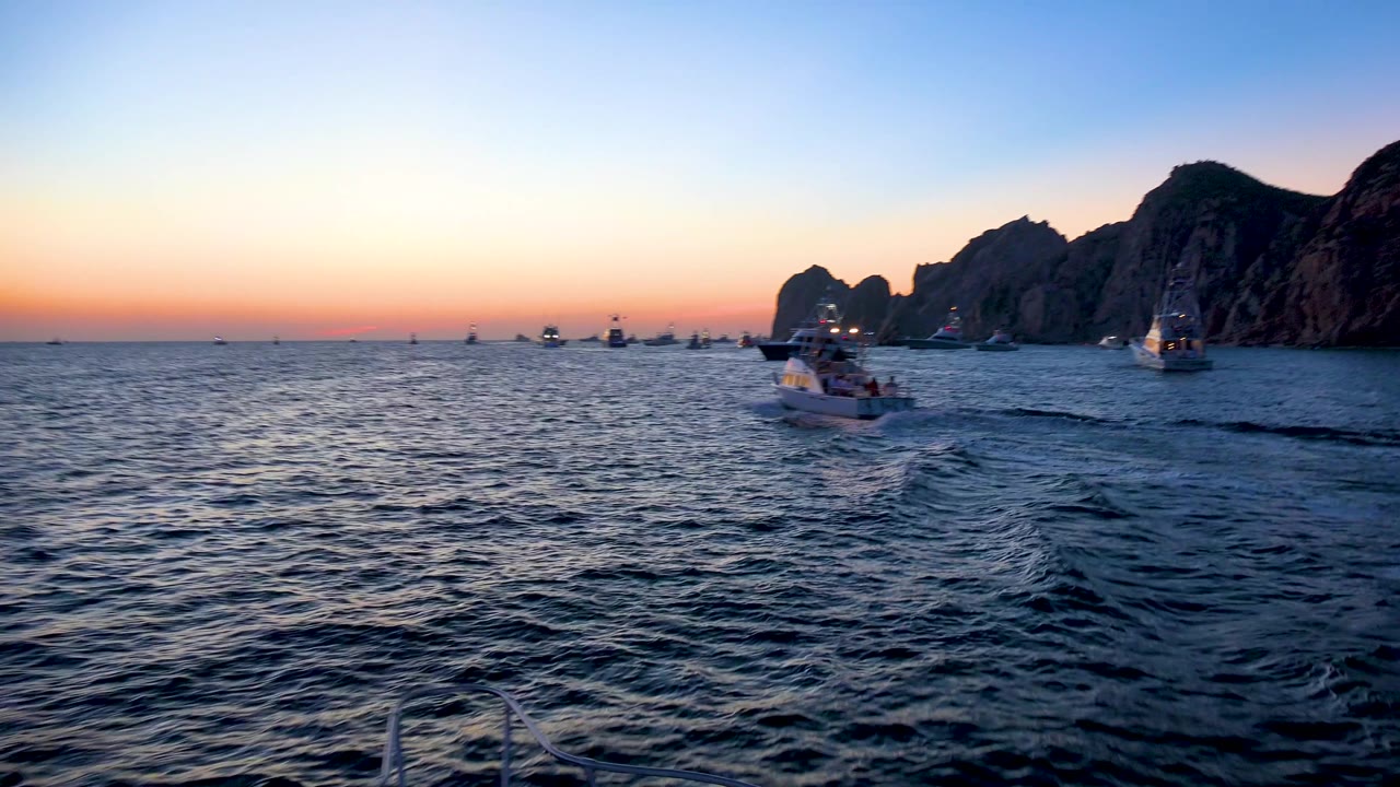 Cabo San Lucas, Mexico dusk seascape with the main boat slightly left of center, many small boats on the horizon, cliffs on the right edge, and no visible people.