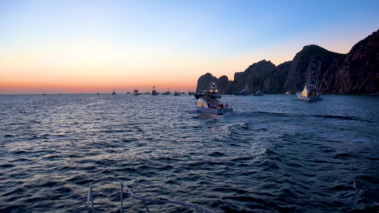 Cabo San Lucas, Mexico frame with the main boat centered lower in the water, distant vessels near the horizon, dark cliff silhouettes to the right, and no visible people.