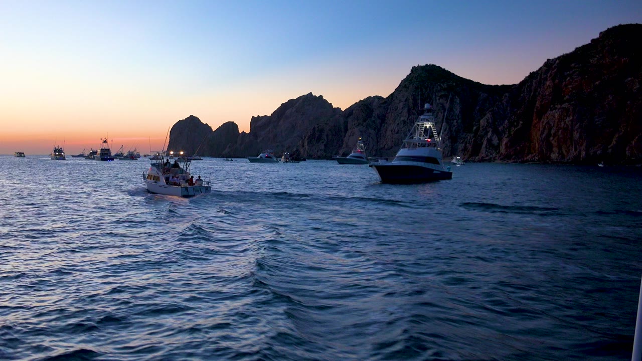 Boats float on choppy water in Cabo San Lucas at dusk, with a small boat left of center, a larger yacht on the right, silhouetted rocks in the background, and no visible people.