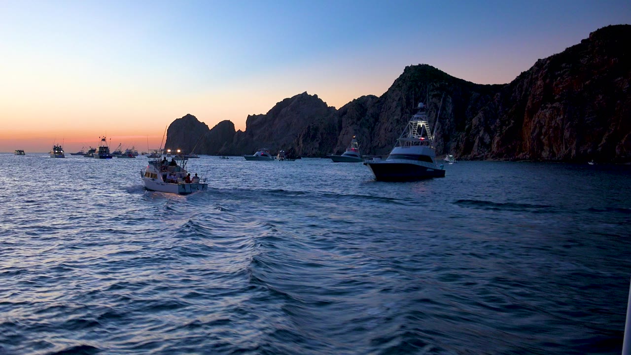A small boat sits nearer the center of the Cabo San Lucas harbor view while a yacht remains to the right, with orange dusk light along the horizon and no visible people.