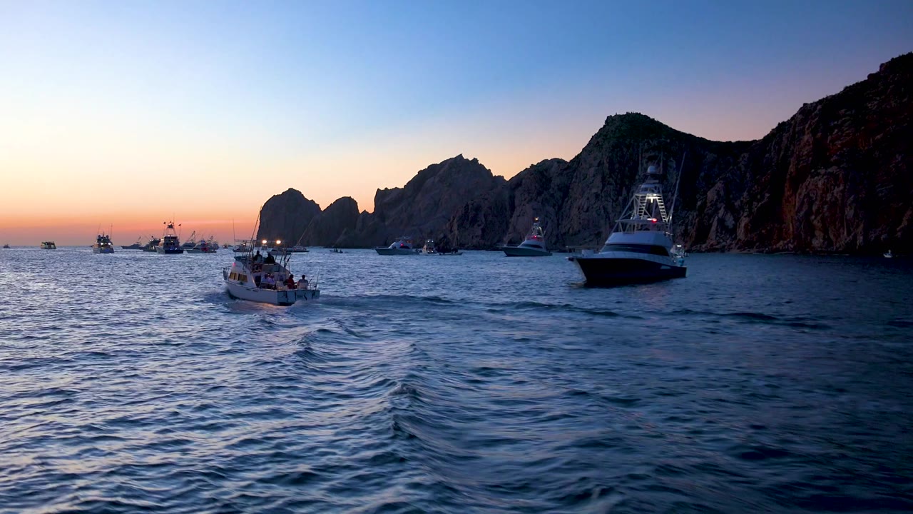 A wider view shows multiple boats across the water in Cabo San Lucas, with the main small boat near center, the larger yacht close to the right edge, rocky cliffs, and no visible people.