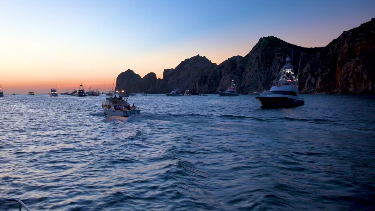 The small boat appears farther left in the Cabo San Lucas dusk scene, with darker choppy water in the foreground, boats scattered near the horizon, rock formations to the right, and no visible people.