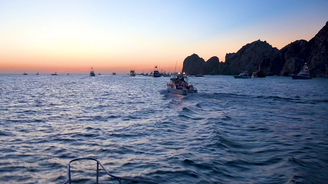 Cabo San Lucas ocean at sunset with a small boat near center, distant boats on the horizon, dark rock formations to the right, foreground boat rail, and no visible people.