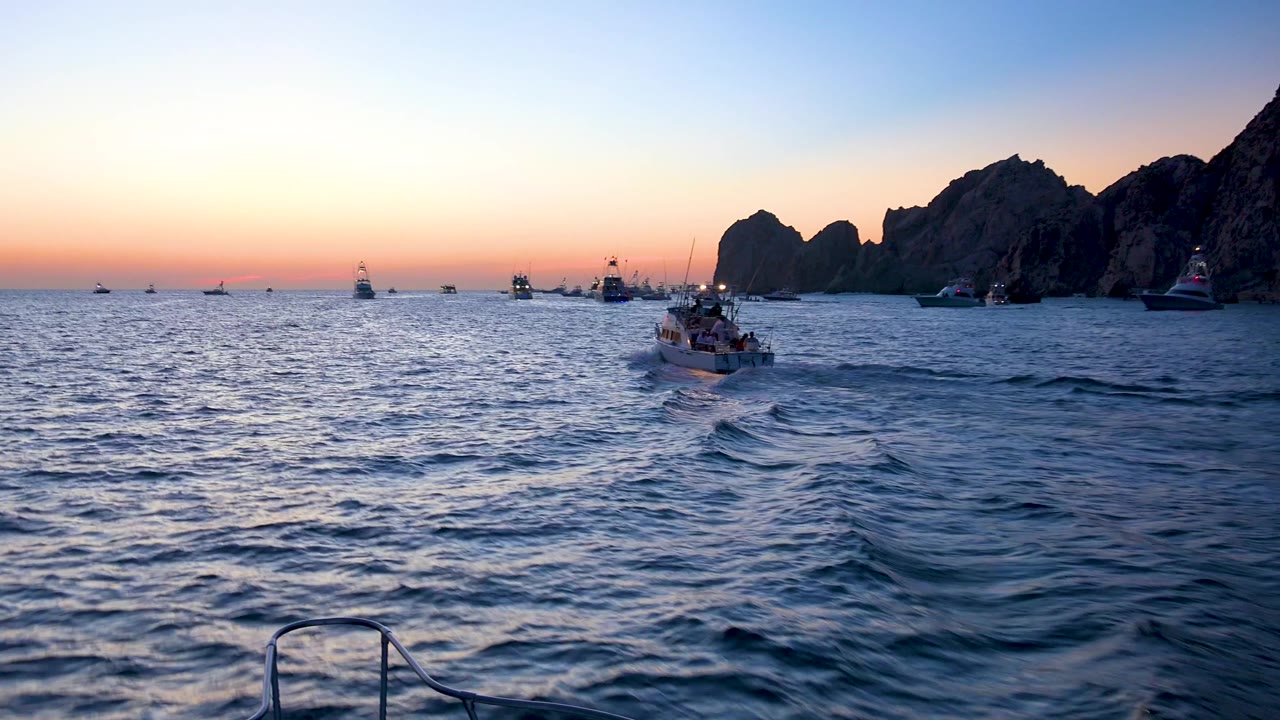 Cabo San Lucas sunset water view with a small boat ahead of the camera, low orange horizon light, several distant boats, right-side rock silhouettes, and no visible people.