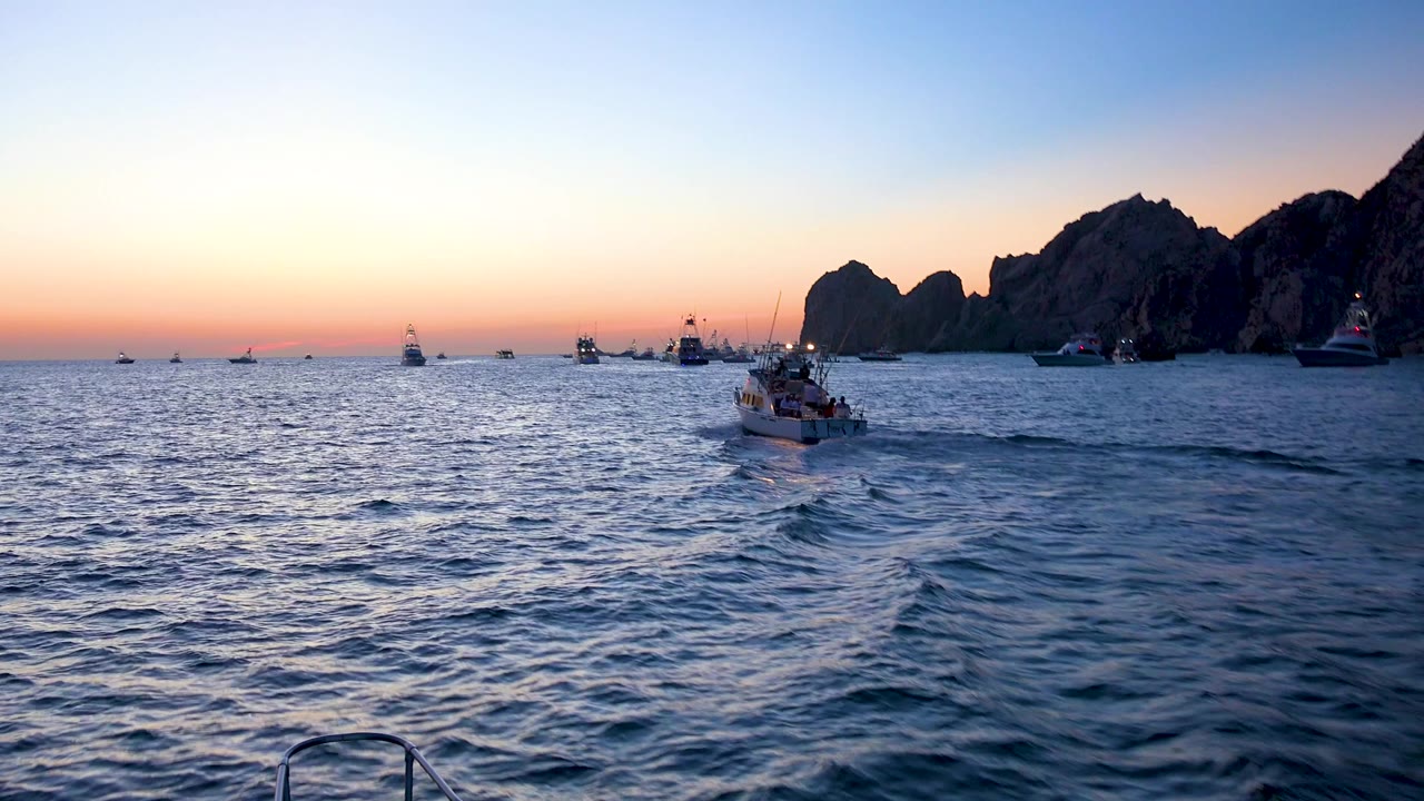 Cabo San Lucas ocean at dusk with a small boat slightly right of center, a pale wake across the water, distant boats, dark shoreline rocks, and no visible people.